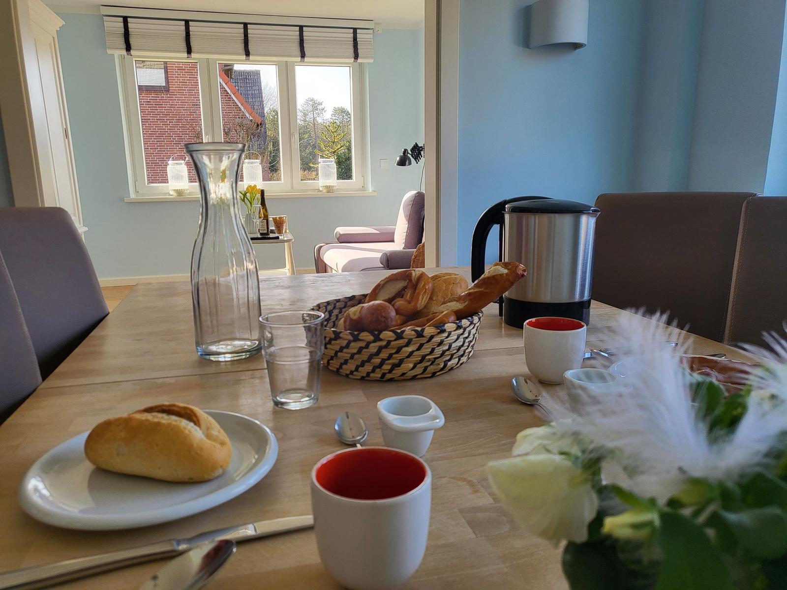 Breakfast table with bread, mugs, and coffee pot on wooden table. Background shows window and seating area.