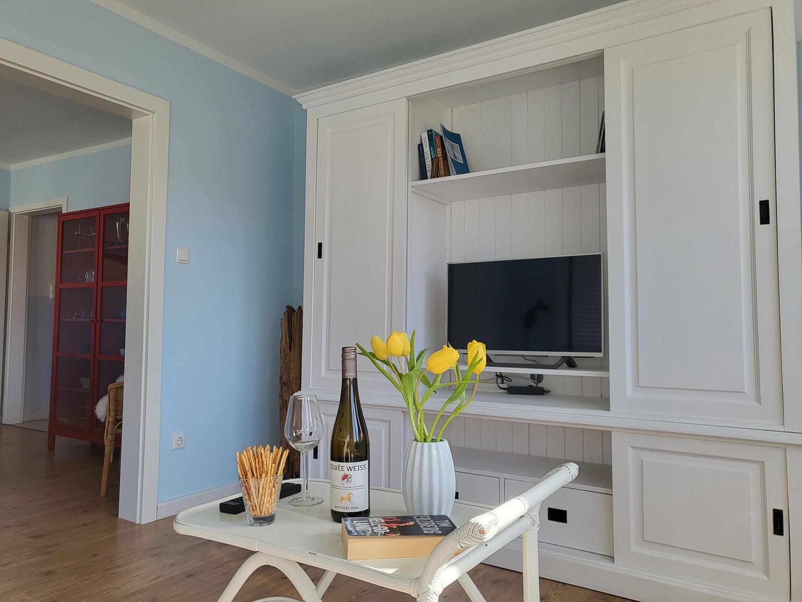 Living room with white cabinet, TV, table with flowers and wine.
