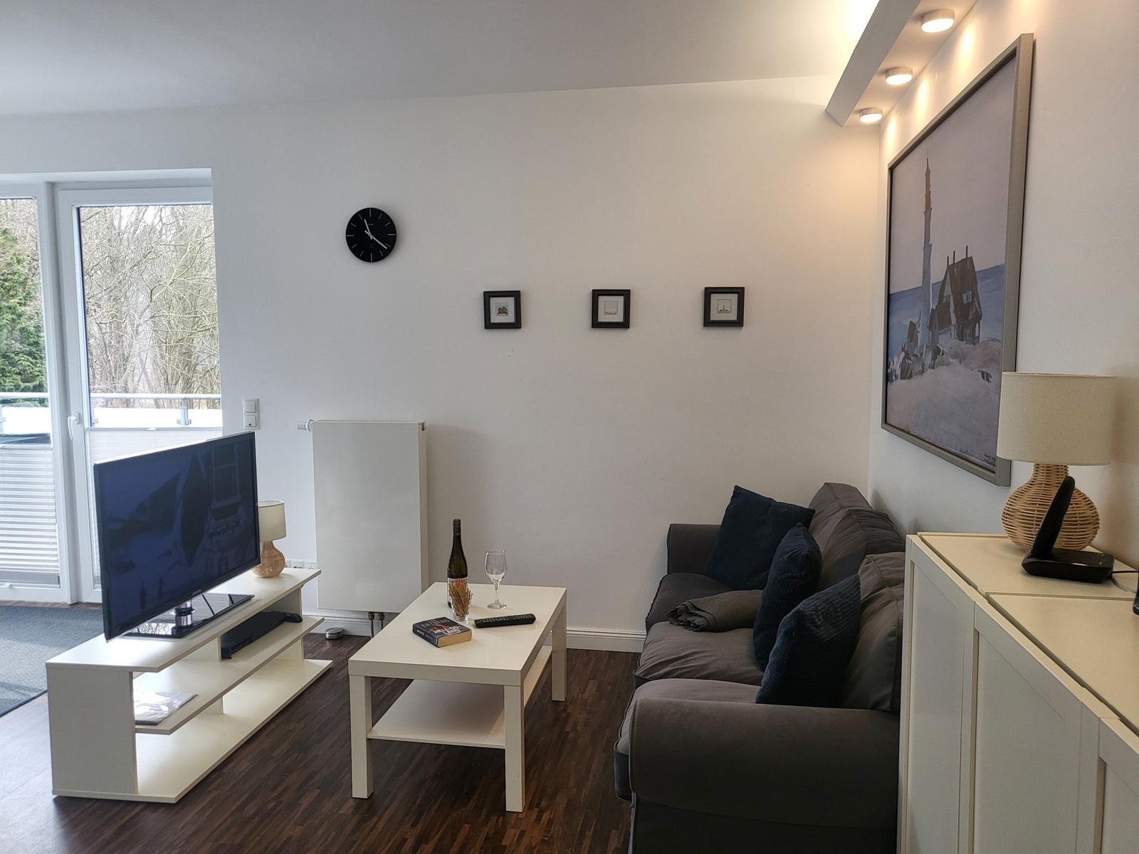 Wohnzimmer mit Sofa, Couchtisch und Fernseher. An der Wand hängen Bilder und eine Uhr.