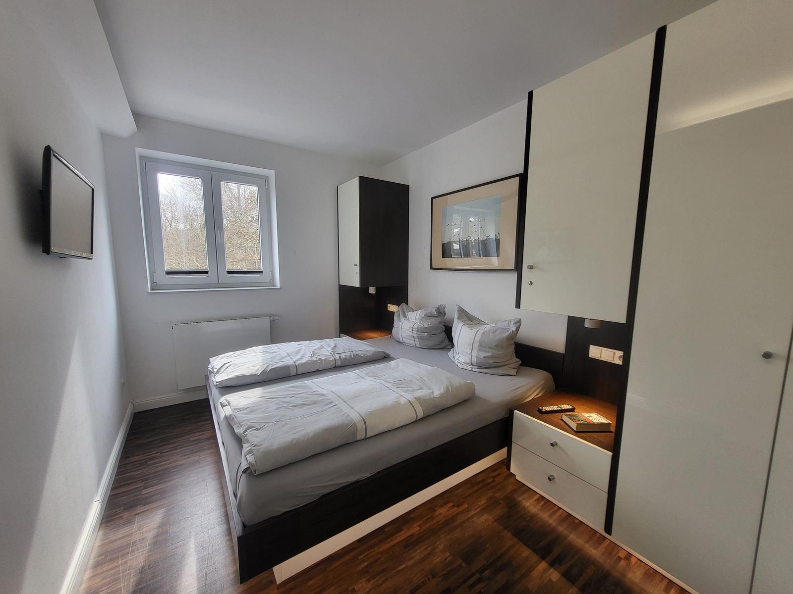 Schlafzimmer mit Doppelbett, Fenster, Kleiderschrank und Fernseher an der Wand.