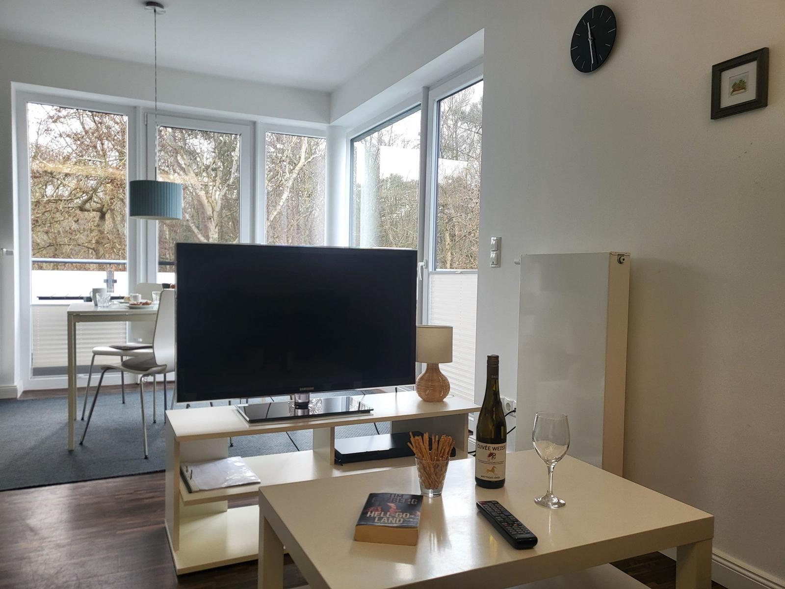 Wohnzimmer mit Fernseher, Tisch und Fenstern. Auf dem Tisch steht eine Weinflasche und ein Glas.