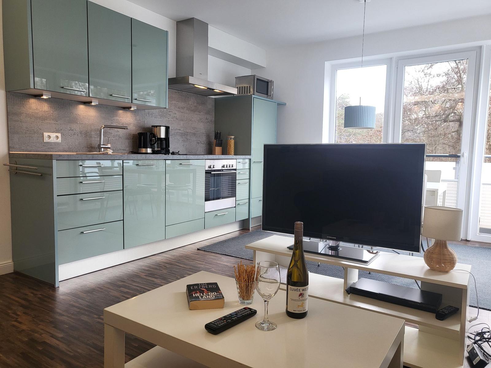 Küche und Wohnzimmer mit TV, Tisch und Fenster zur Terrasse.