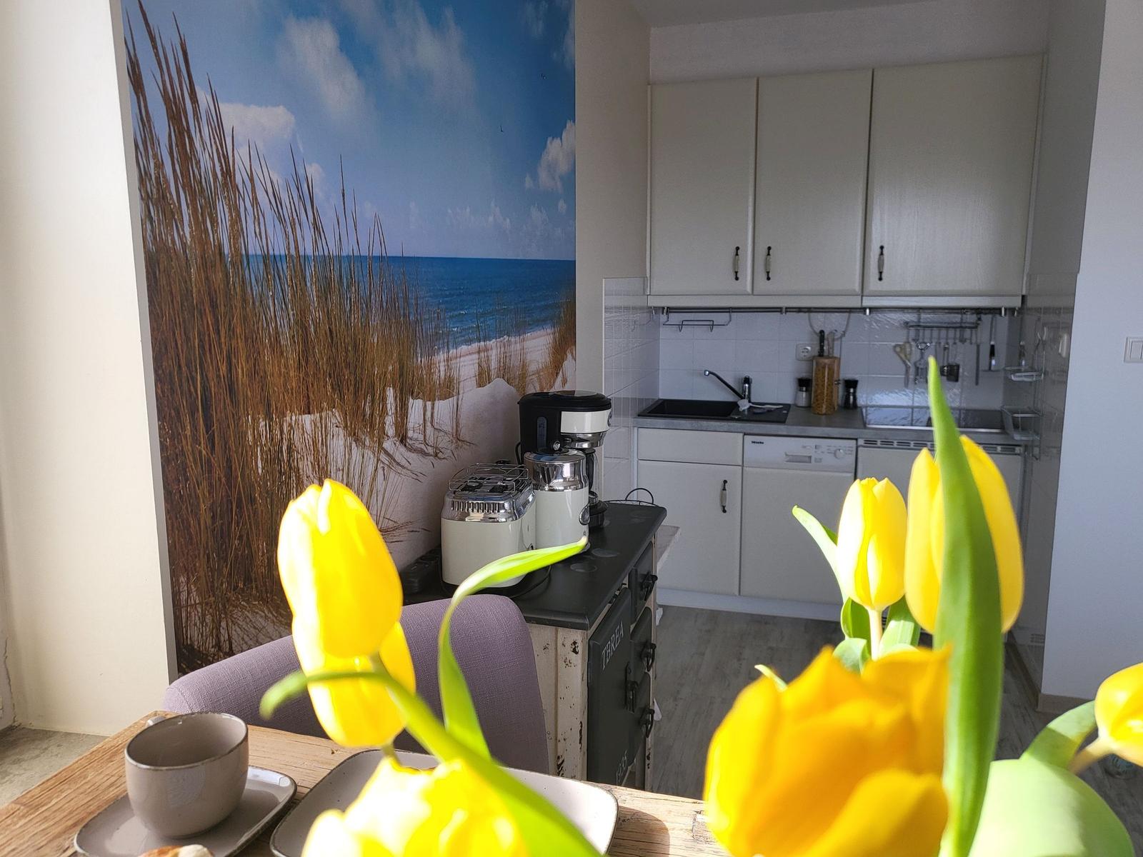 Küche mit Küchenschränken und Küchenzeile. Wandbild zeigt Strand. Vordergrund: gelbe Tulpen.