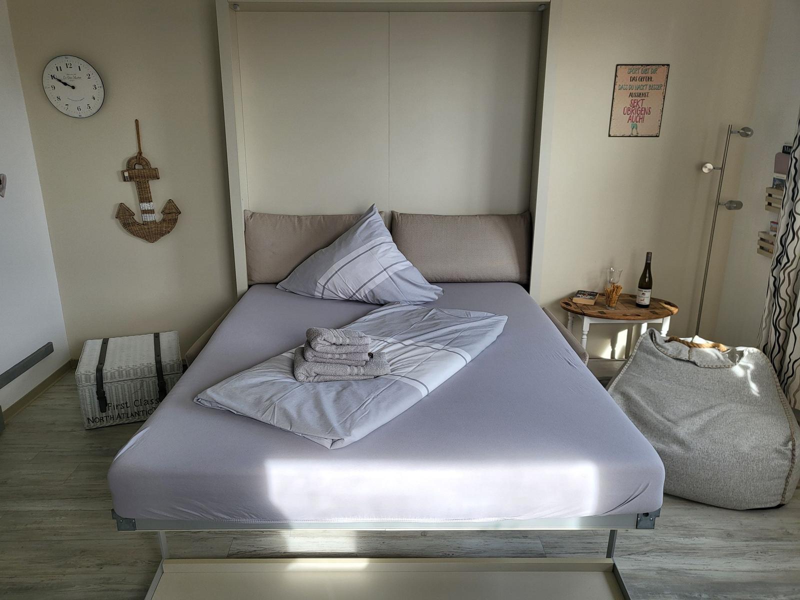 Ein Bett mit Laken und Kissen, Wanduhr, Ankerdekoration und Sitzkissen.