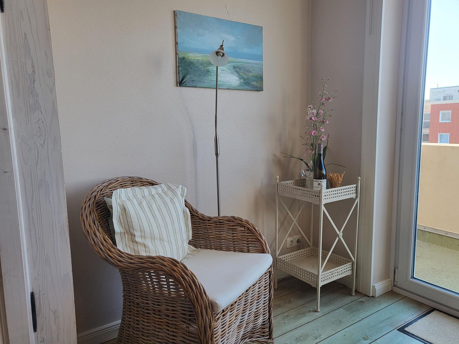 Wickerstuhl mit Kissen, Wandbild, Stehlampe und kleiner Tisch mit Blumen und Weinflasche.