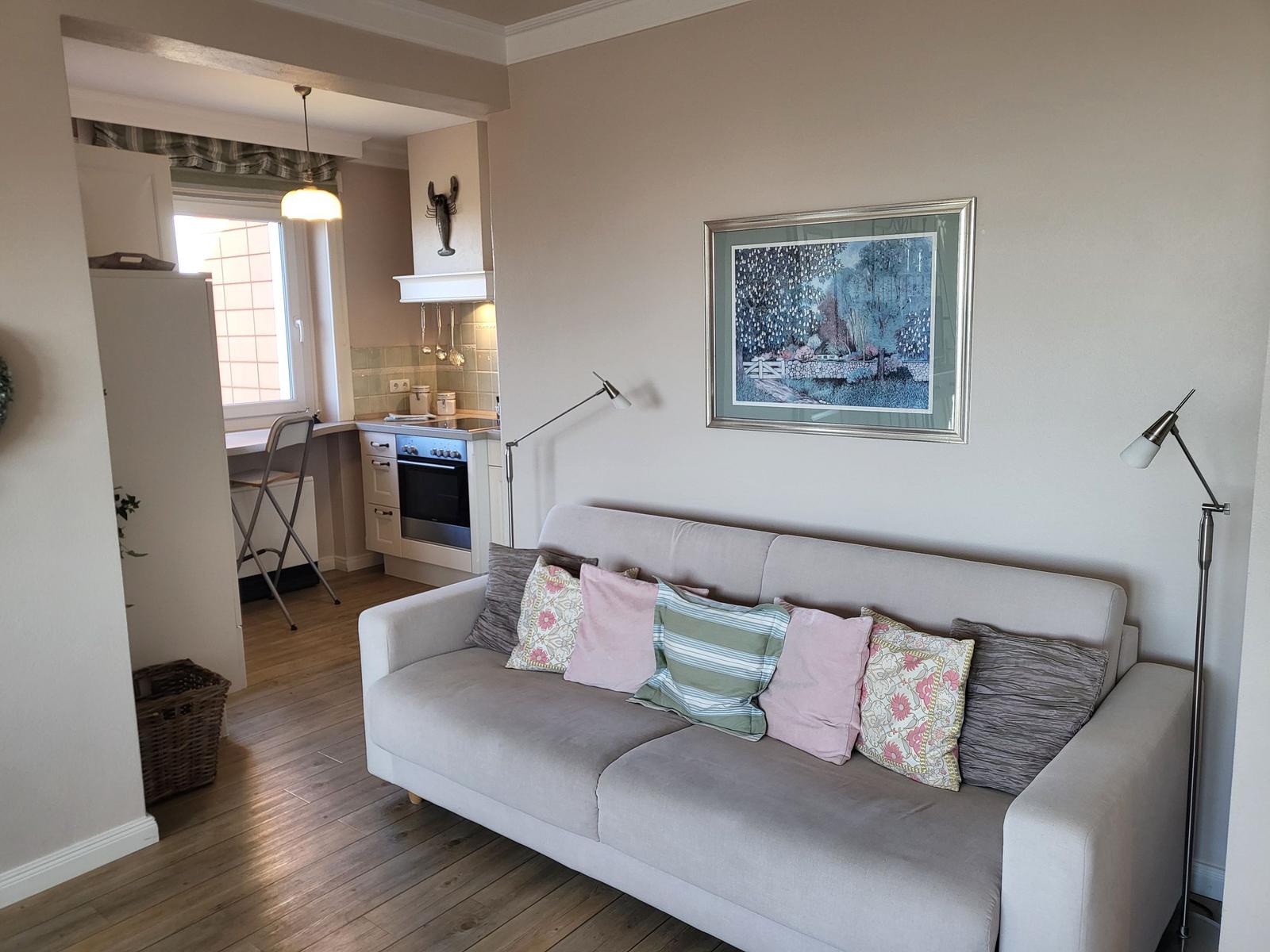 Ein Wohnzimmer mit Sofa, Kissen und Wandbild. Küche ist im Hintergrund sichtbar.
