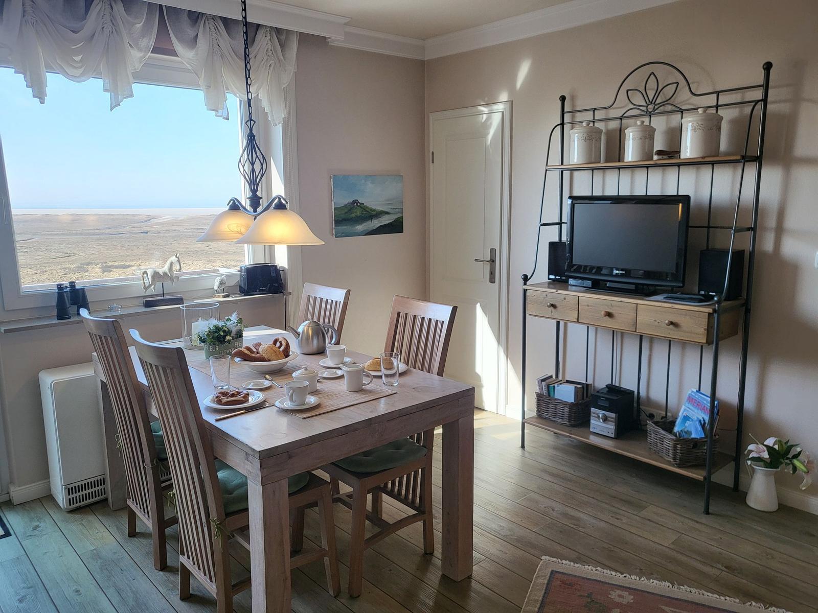 Esszimmer mit Holztisch, Stühlen und Fernseher. Großer Fensterblick auf Landschaft.