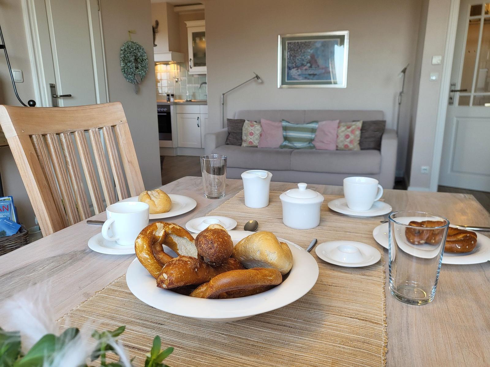 Frühstückstisch mit Brot, Kaffeetassen und Glas. Im Hintergrund Sofa und Küche.