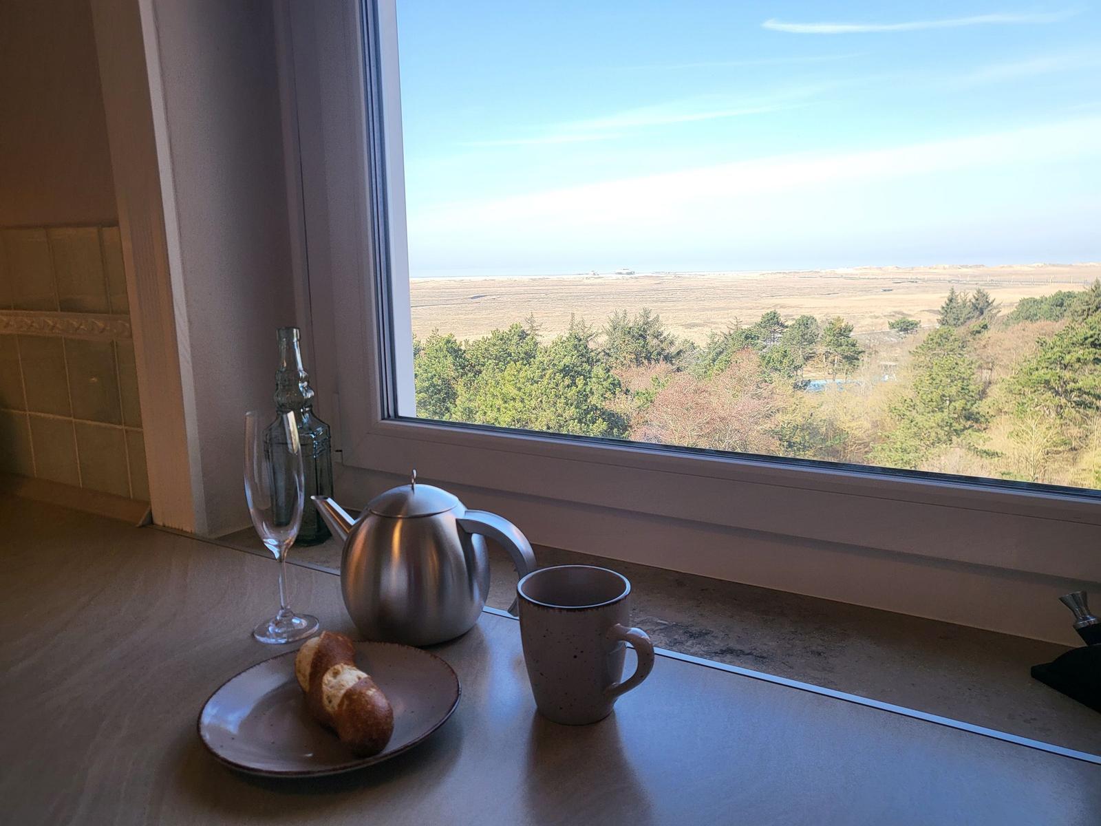 Auf dem Fensterbrett steht eine Tasse, Kanne und ein Brötchen. Draußen ist eine Landschaft mit Bäumen und Feldern.