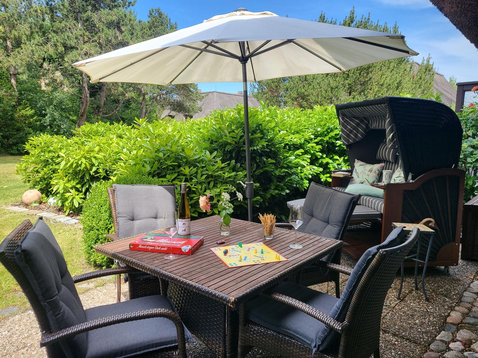 Terrasse mit Tisch, Stühlen, Sonnenschirm und Liegestuhl. Auf dem Tisch liegt ein Spiel.