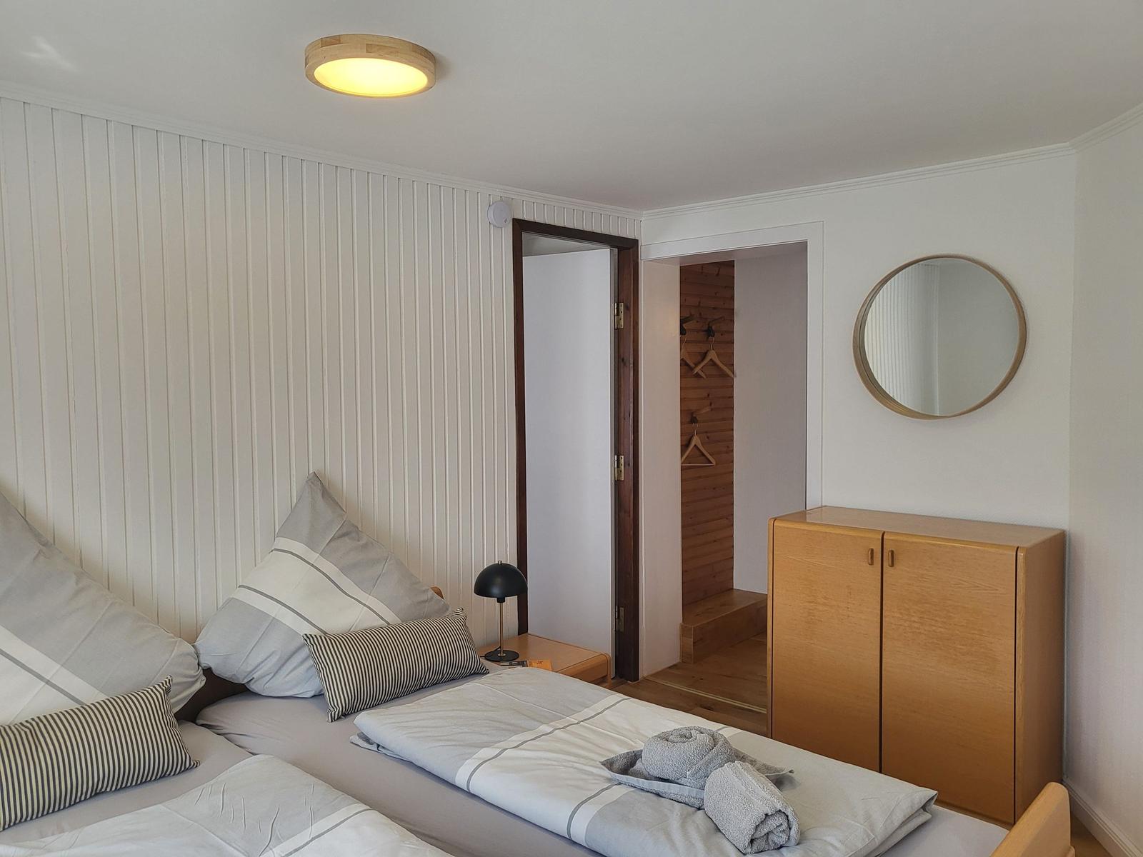 Schlafzimmer mit Bett, Kleiderschrank und Spiegel. Tür führt in einen Flur mit Kleiderhaken.