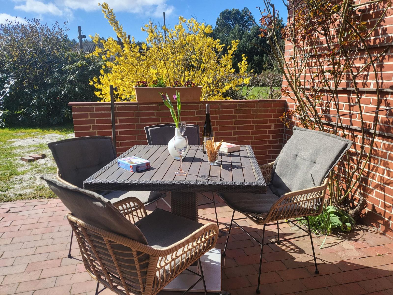 Terrasse mit Tisch und vier Stühlen, Blumen und Weinflasche darauf, umgeben von Ziegelmauer und Grünflächen.