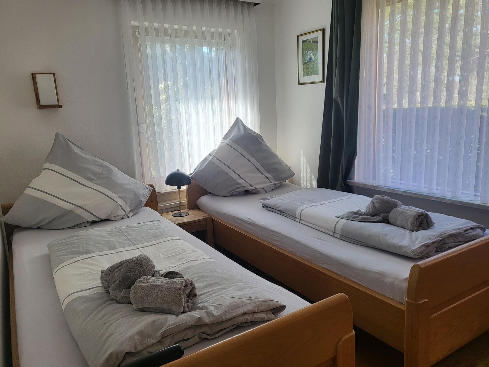 Zwei Einzelbetten mit Bettwäsche und Handtüchern. Fenster mit Jalousien und Vorhängen.