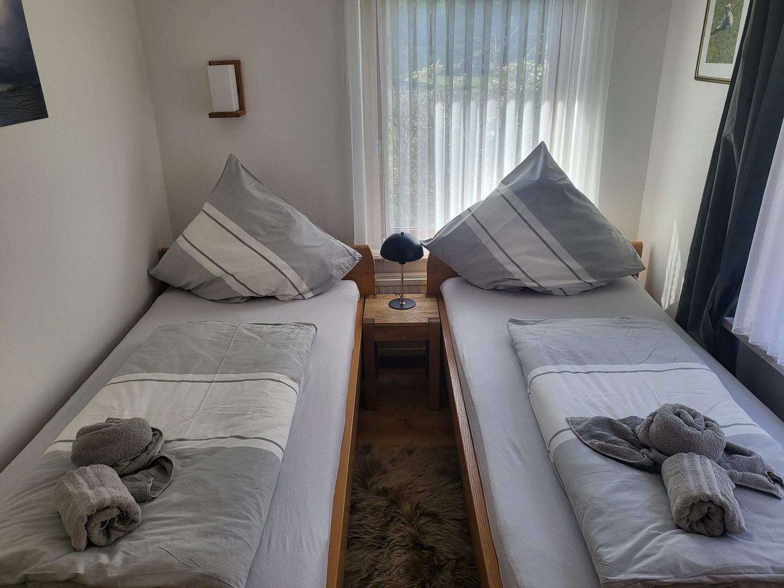 Zwei Einzelbetten mit grauen Bettwäschen und Handtüchern. Zwischen den Betten steht ein Nachttisch mit Lampe.