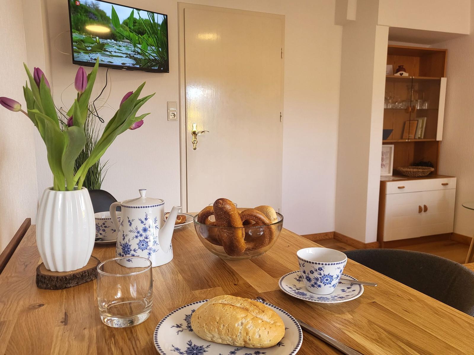 Esstisch mit Kaffeetasse, Brötchen und Blumen. Fernseher an der Wand.