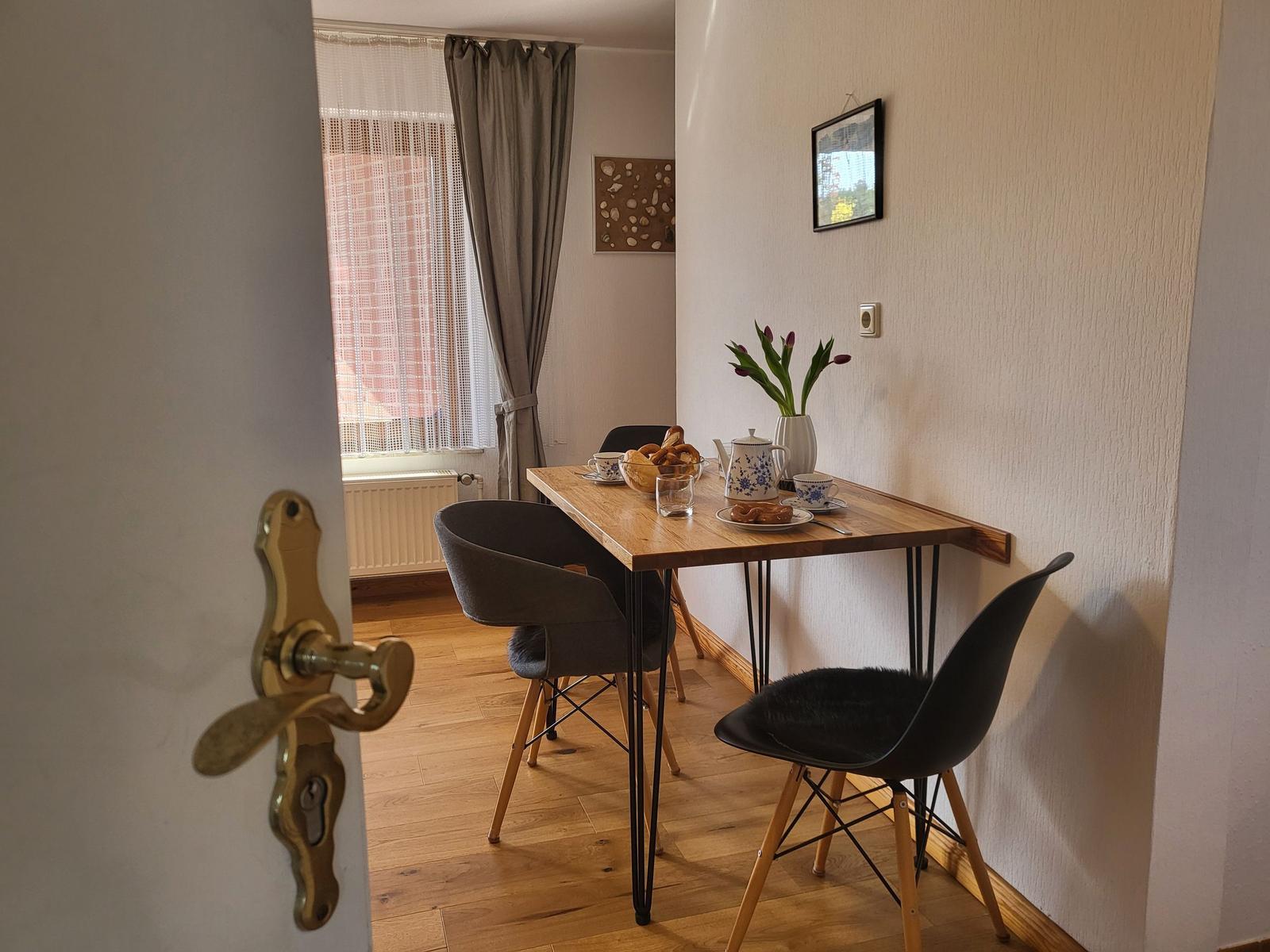 Essbereich mit Tisch, Stühlen und Blumen. Fenster mit Vorhängen und Wandbild.