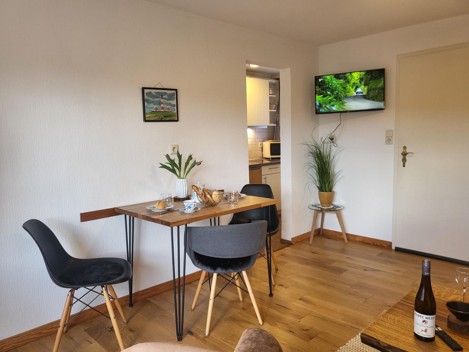 Essbereich mit Tisch, Stühlen und Fernseher an der Wand. Küche im Hintergrund sichtbar.