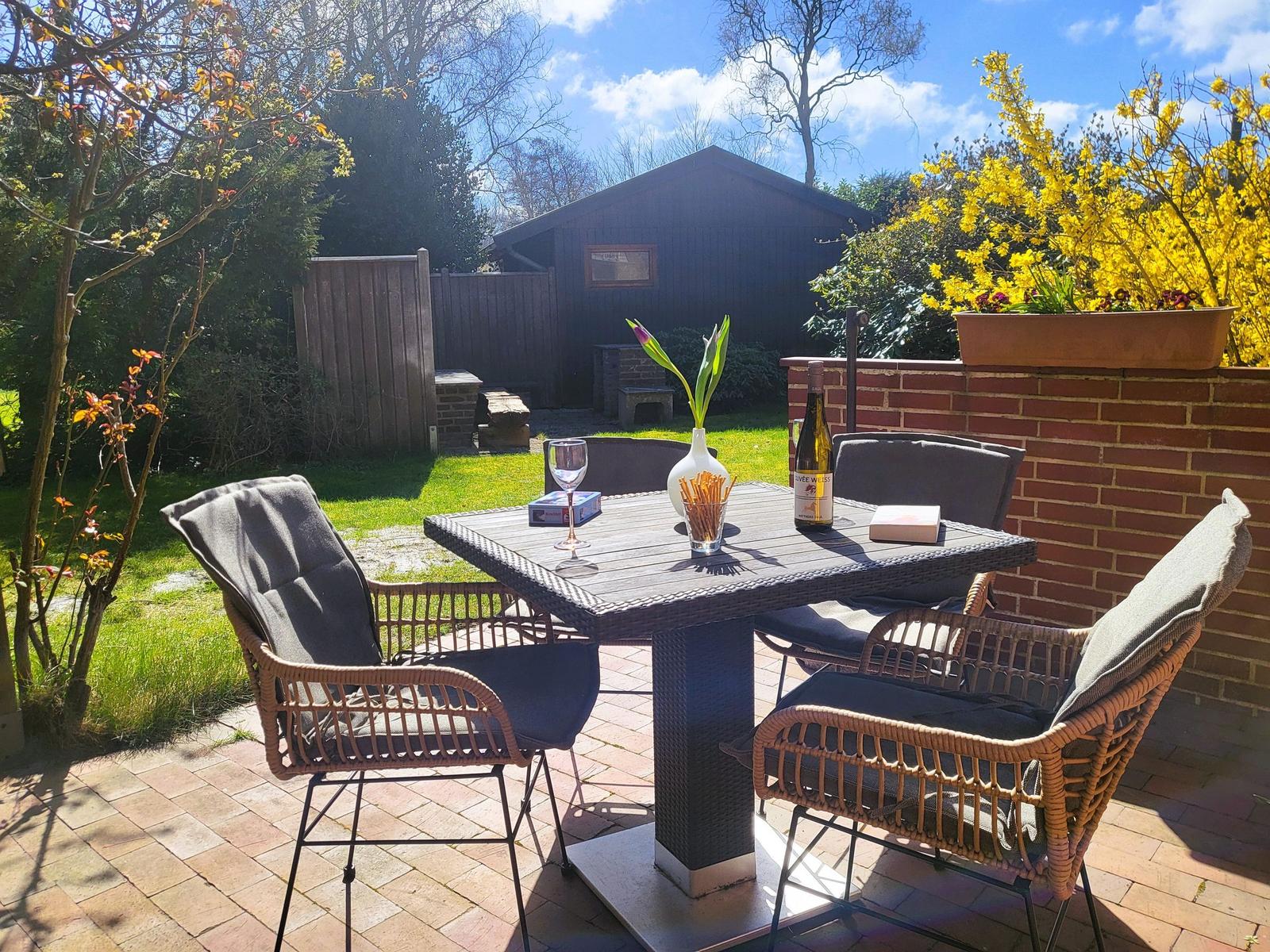 Terrasse mit Tisch und Stühlen, Wein und Blumen auf dem Tisch, Hintergrund mit Garten und Holzhaus.