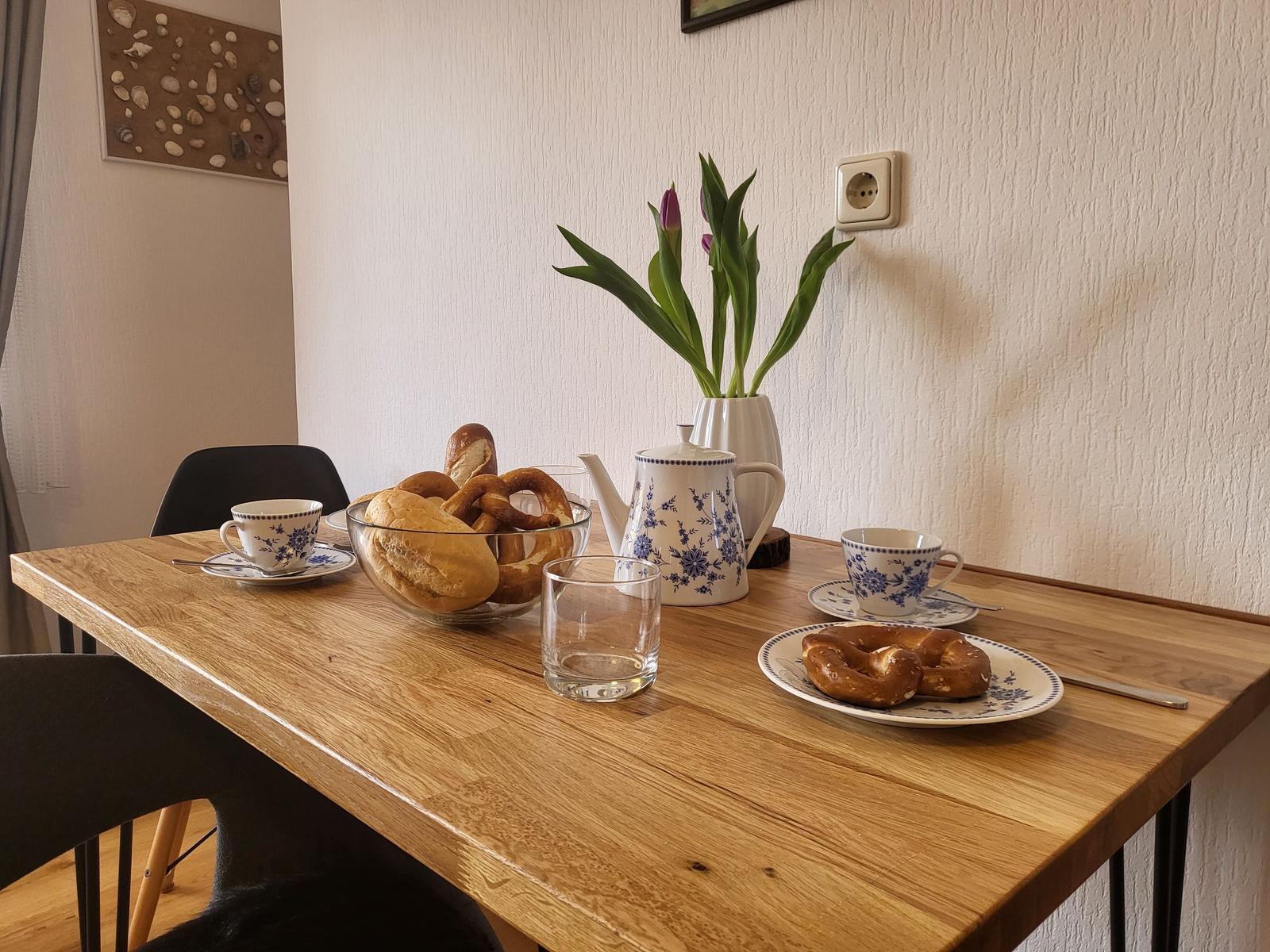 Holztisch mit Tassen, Kaffeekanne, Brötchen und Tulpenblüten.
