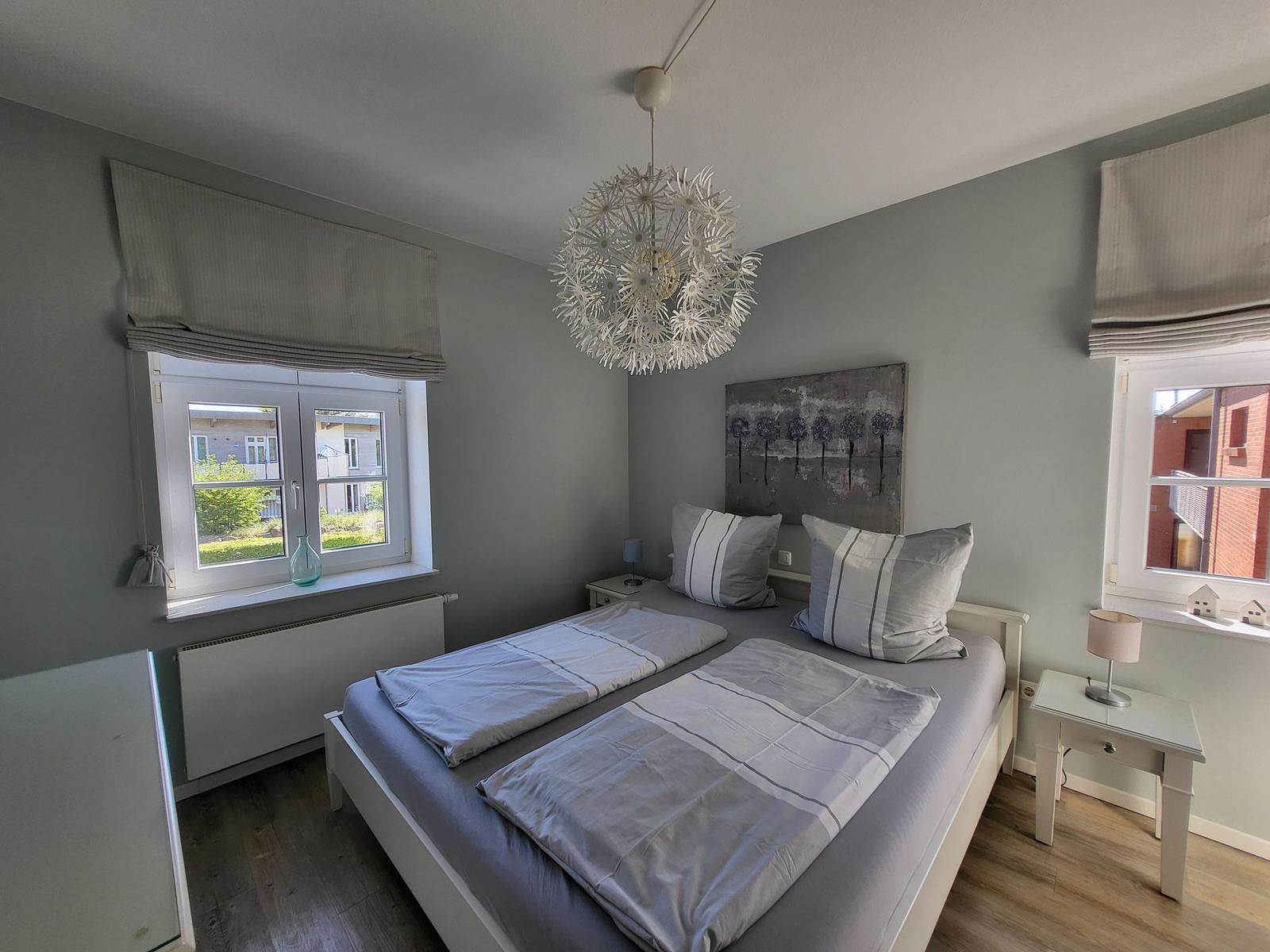 Schlafzimmer mit Doppelbett, zwei Fenstern und Deckenleuchte.