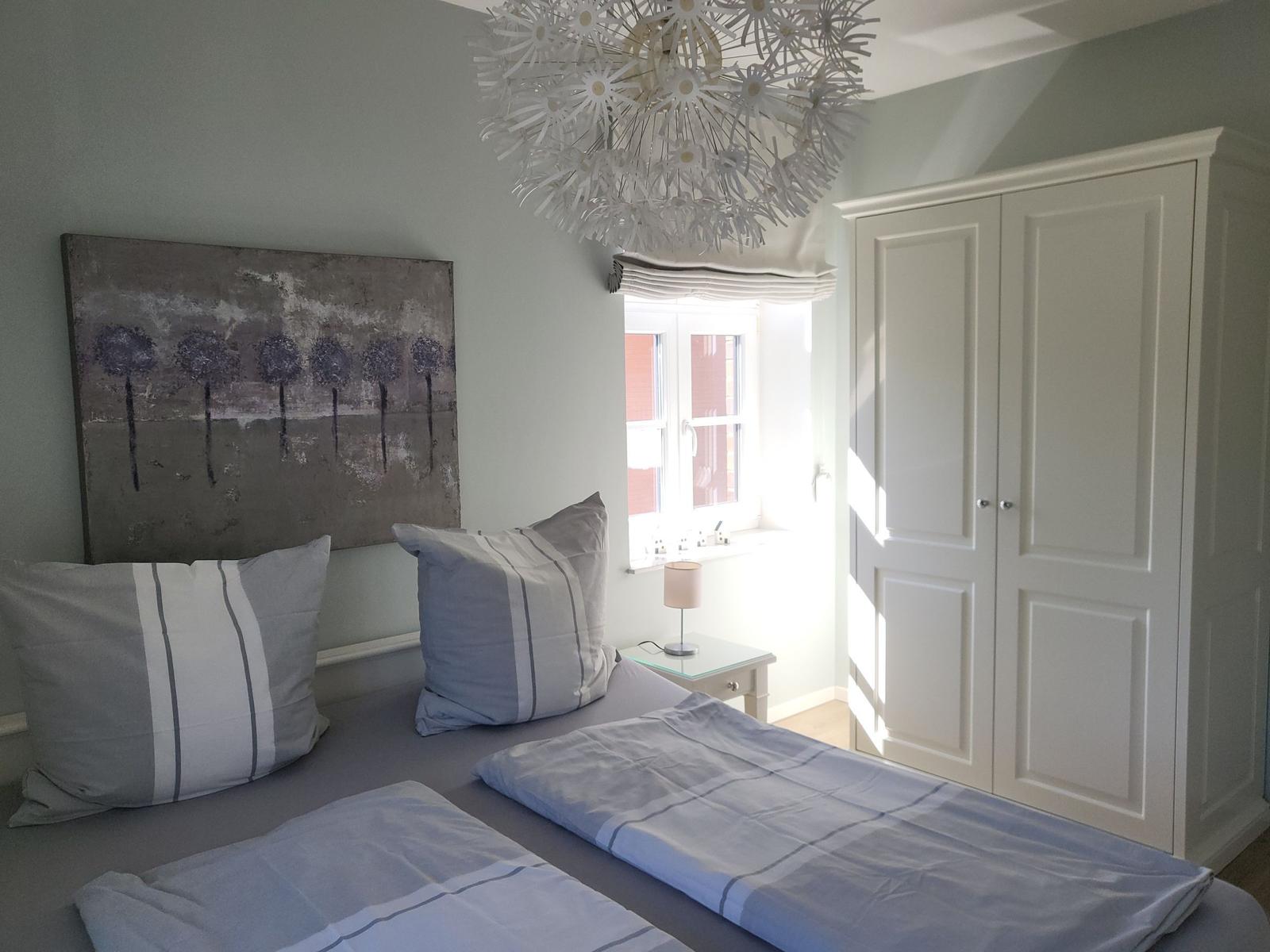 Schlafzimmer mit Doppelbett, Kleiderschrank und Fenster. Deckenleuchte und Wandbild sind sichtbar.