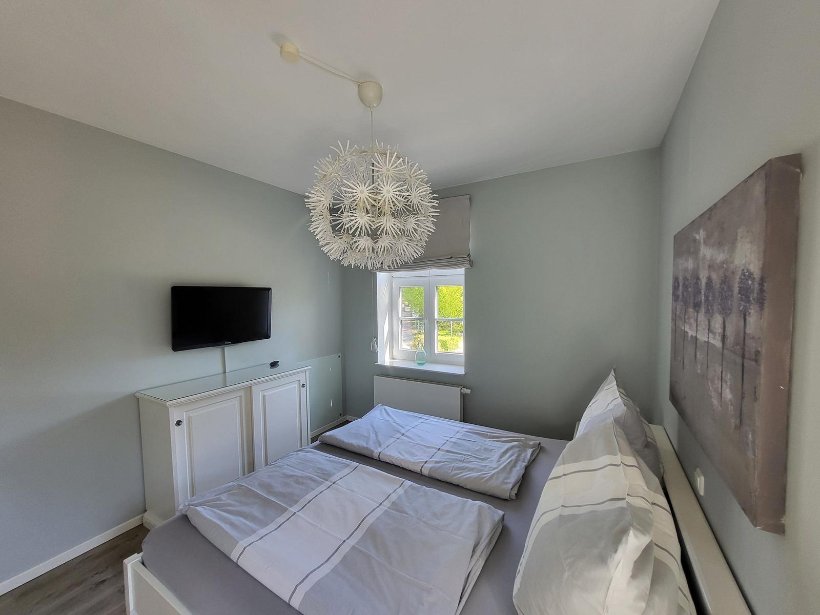 Schlafzimmer mit Doppelbett, Fenster, Fernseher und Deckenleuchte.