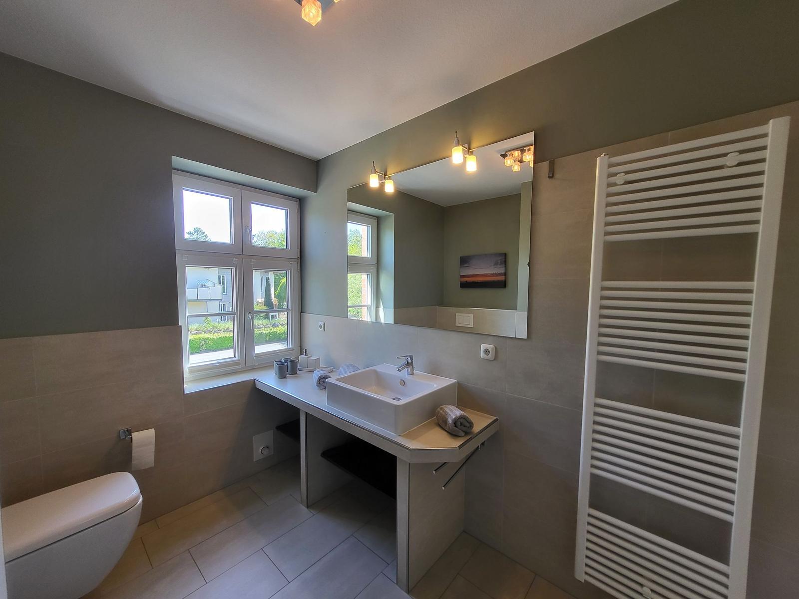 Badezimmer mit Waschtisch, Spiegel, Fenster und Heizkörper.