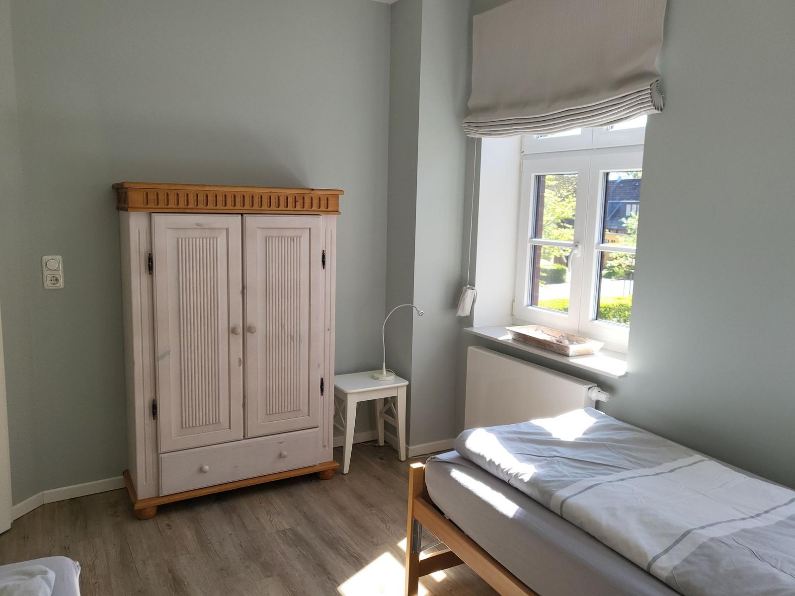 Ein Schlafzimmer mit Bett, Kleiderschrank und Fenster. Holzboden und hellgraue Wände.