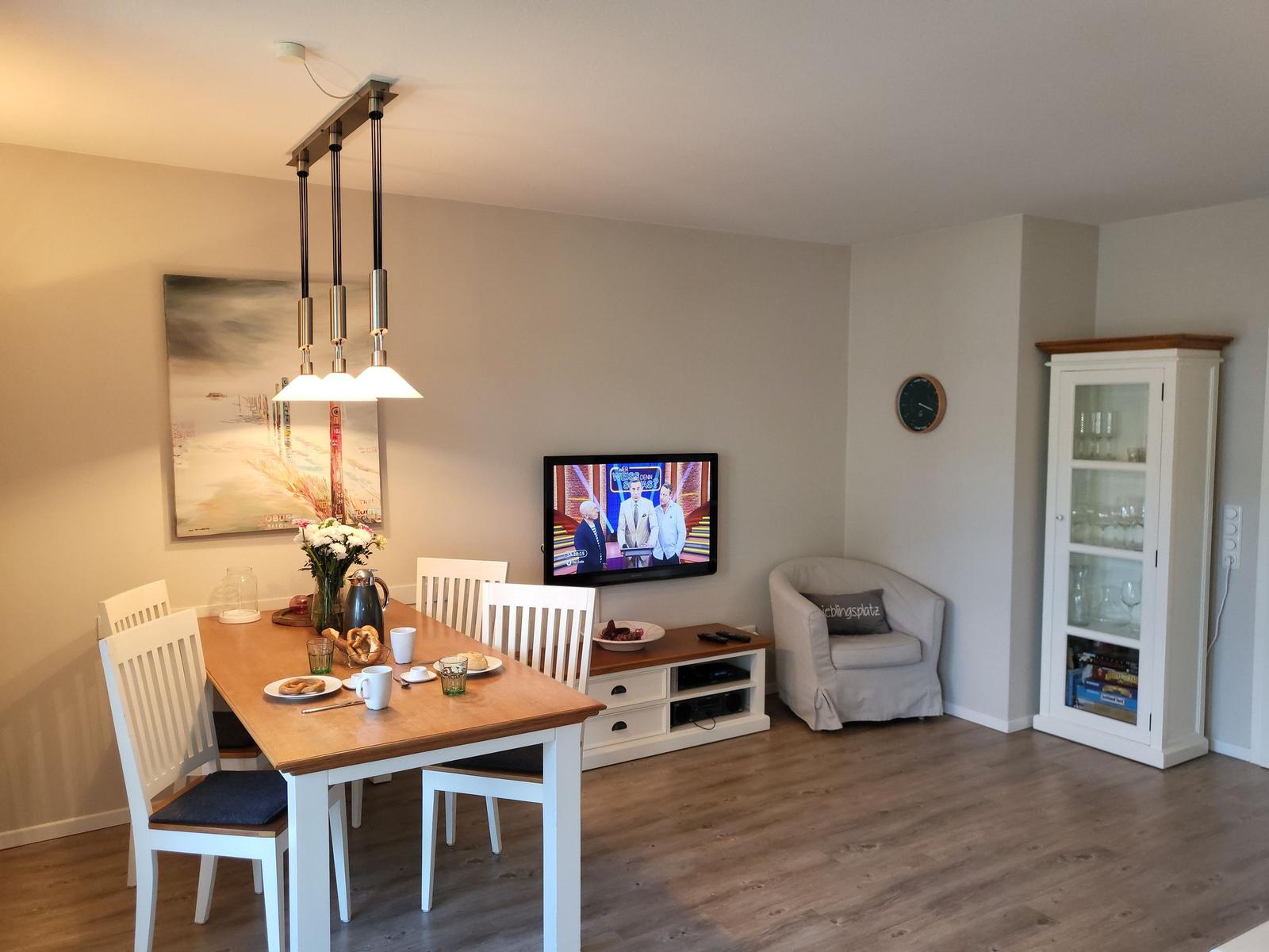 Essbereich mit Tisch, Stühlen und Fernseher. Neben einem Sessel steht ein Glasvitrinenschrank.