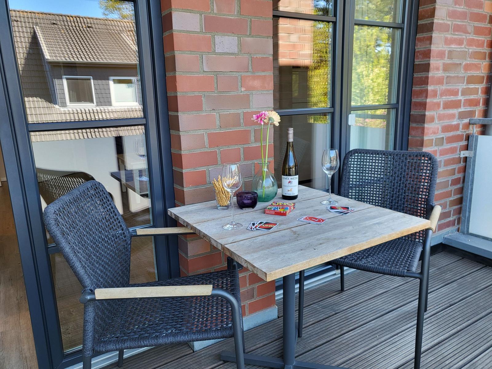 Terrasse mit Tisch, zwei Stühlen, Weinflasche und Blumen. Holzdecke und Backsteinmauer.