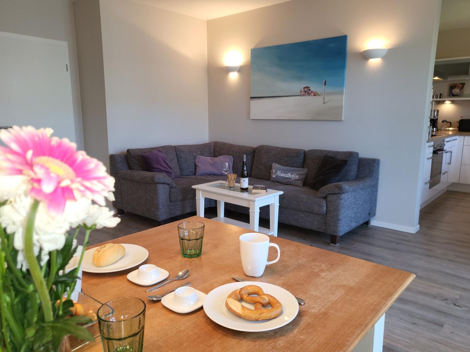 Essbereich mit Tisch, Kaffeetasse und Brötchen. Im Hintergrund Wohnzimmer mit Sofa und Wandbild.