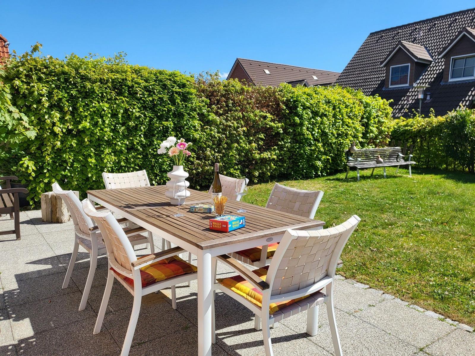 Terrasse mit Tisch und Stühlen, Blumen, Wein und Spielzeug. Hinterhof mit Rasen und Hecke.