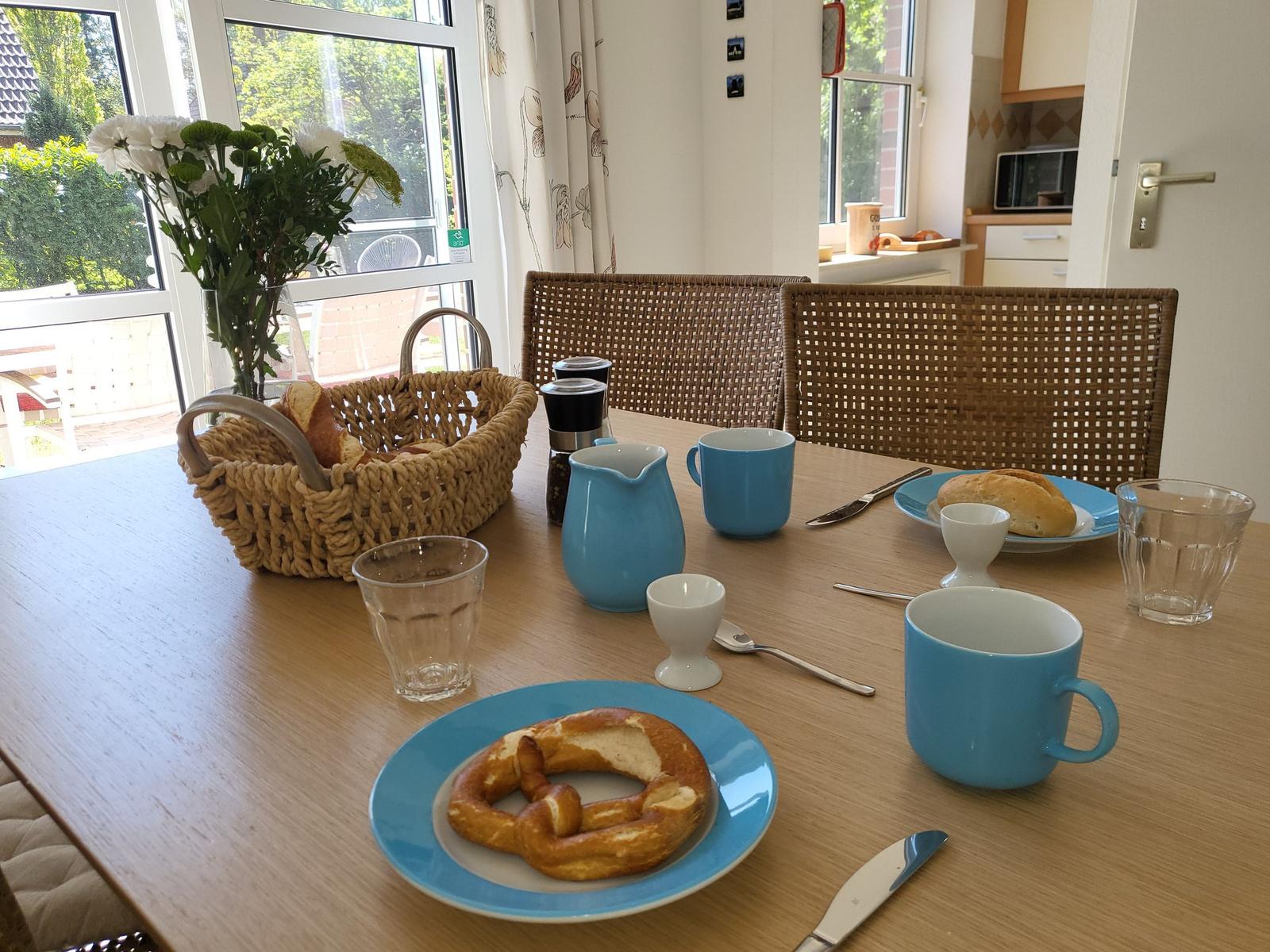 Frühstückstablett mit Brotkorb, Blumen, Tassen und einem Brezel auf einem Holztisch.