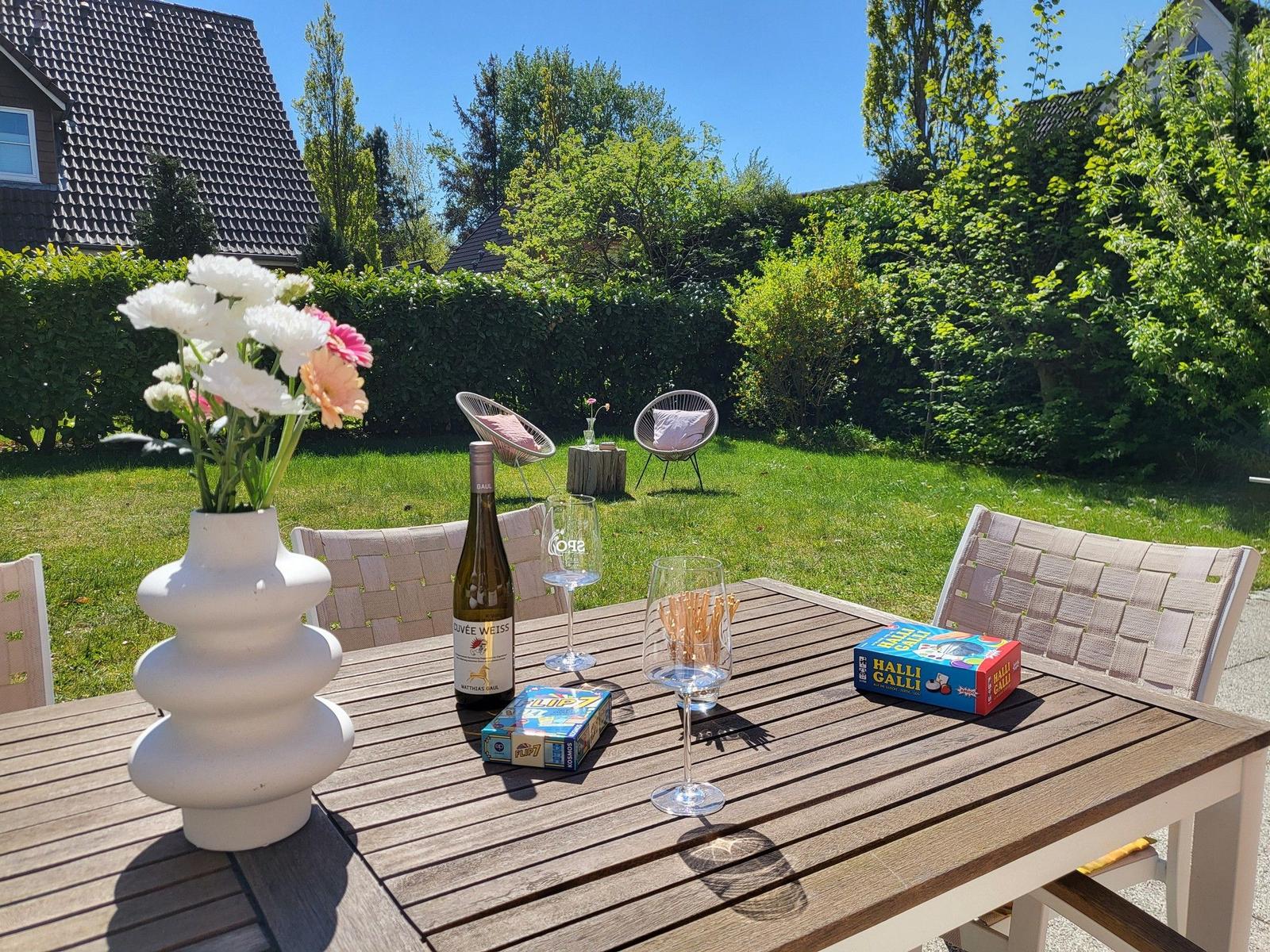 Hochwertiger Holztisch mit Stühlen, Blumen, Wein und Spielzeug im Garten.