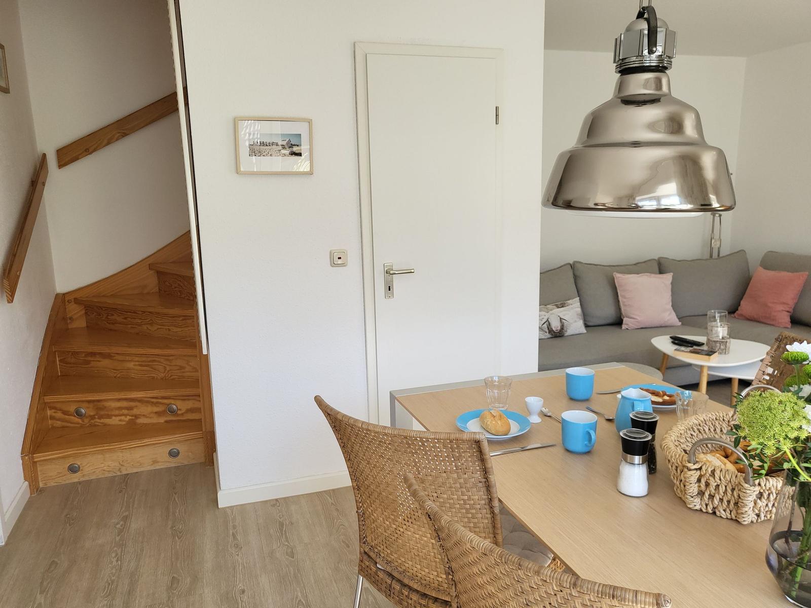 Essbereich mit Tisch, Stühlen und Korb. Im Hintergrund Wohnzimmer mit Sofa und Treppe.