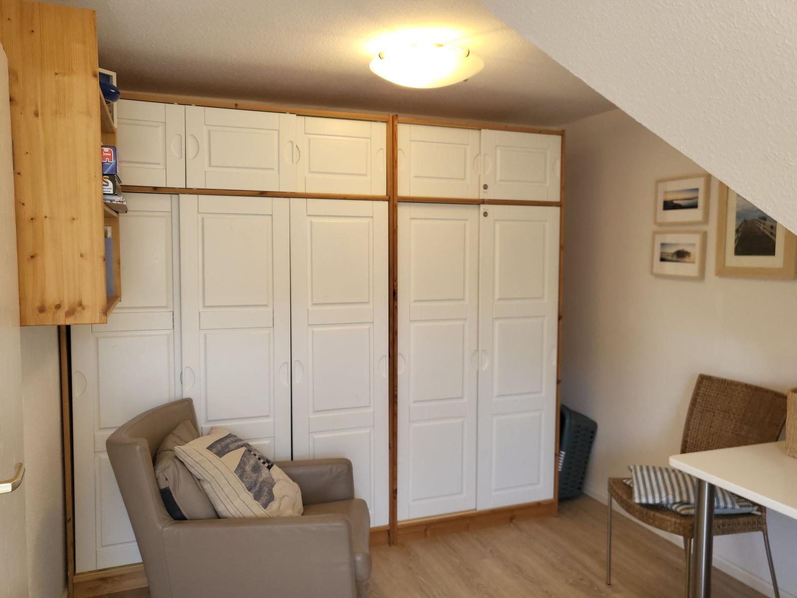 Zimmer mit Kleiderschrank, Sessel und Tisch. Decke geneigt, Wand mit Bildern.