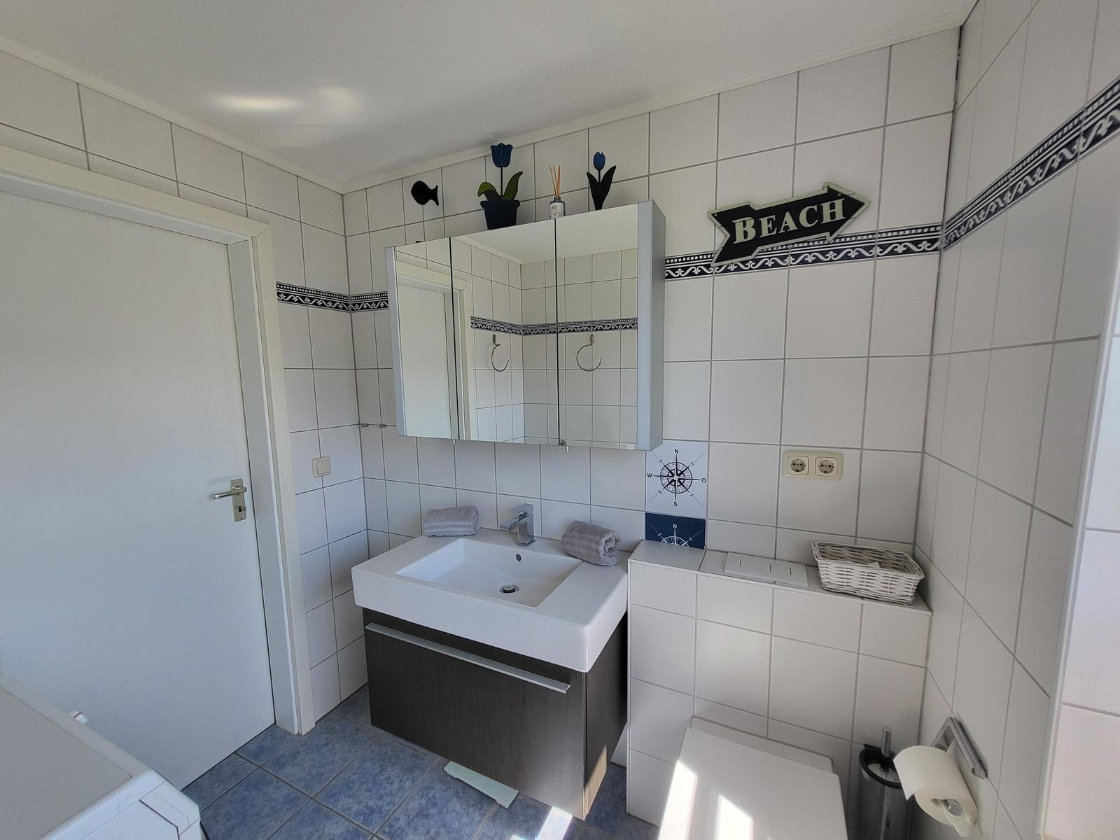 Badezimmer mit Waschbecken, Spiegelschrank und Toilette. Wandverkleidung mit Strandmotiv.