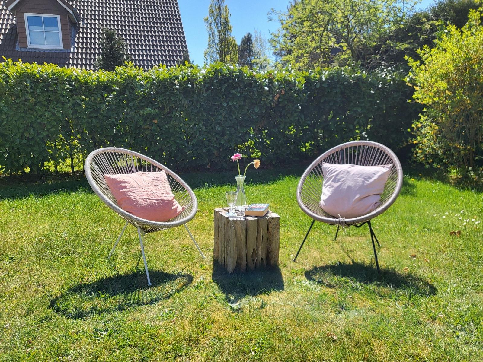 Zwei runde Sessel mit Kissen stehen auf einer Wiese. Dazwischen steht ein Tisch mit Blumen und einem Buch.