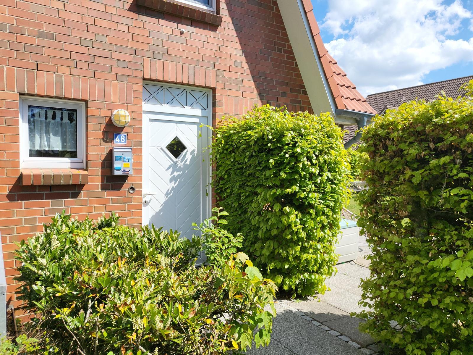 Haus mit rotem Backsteinfassade, weißer Tür und grünen Sträuchern im Vorgarten.