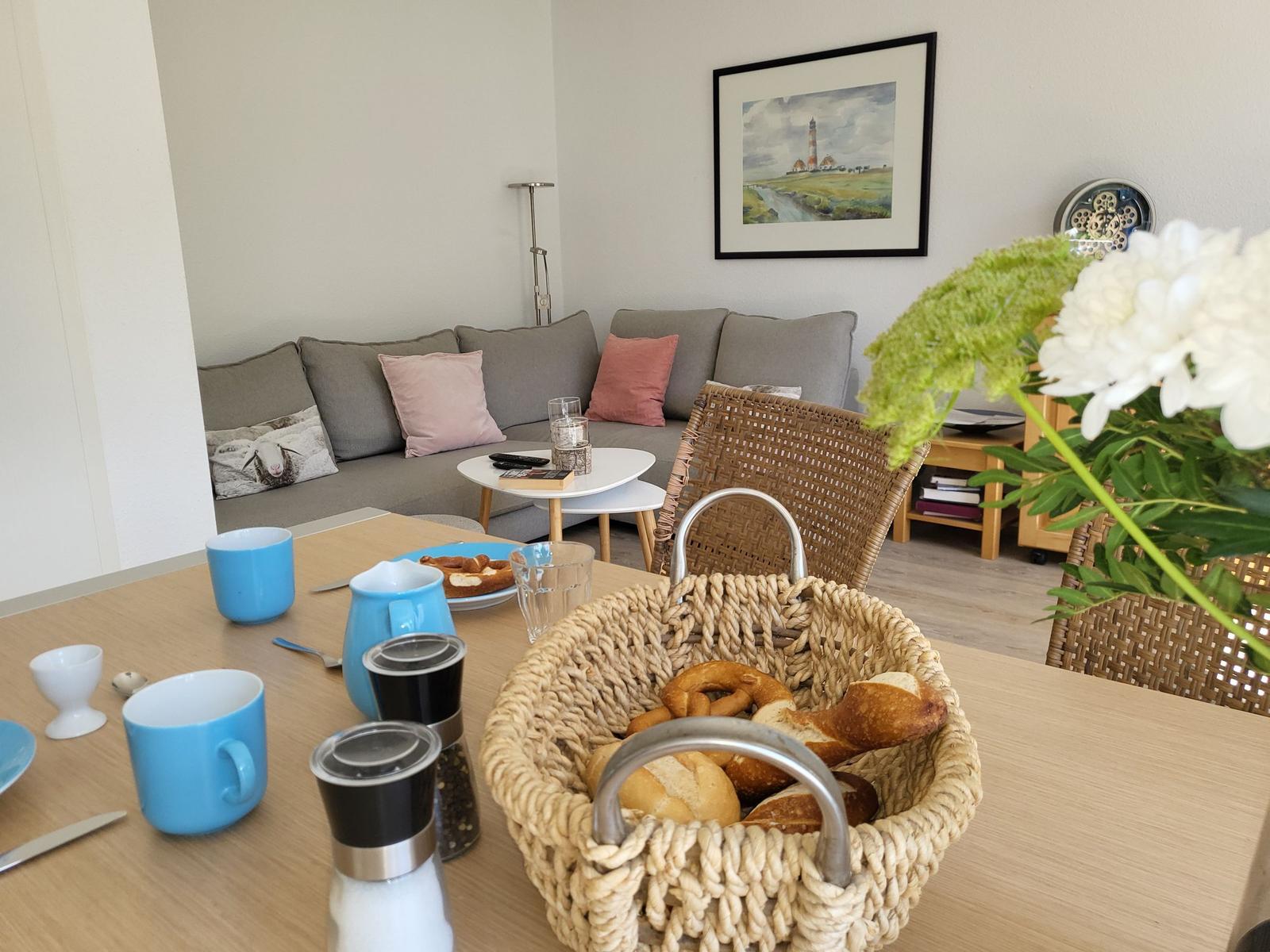 Esszimmer mit Tisch, Stühlen und Korb mit Brot. Im Hintergrund Sofa und Wandbild.