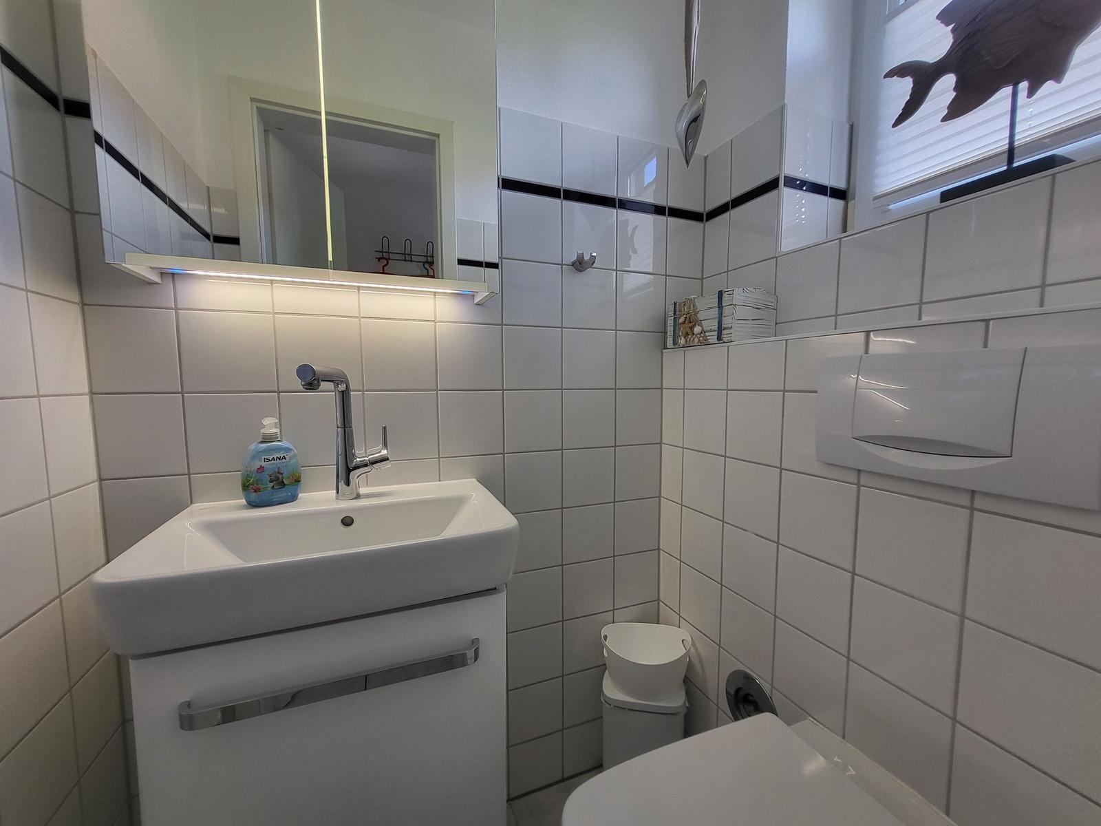 Badezimmer mit Waschbecken, Spiegel und Toilette. Weiße Fliesen und Fenster mit Jalousie.