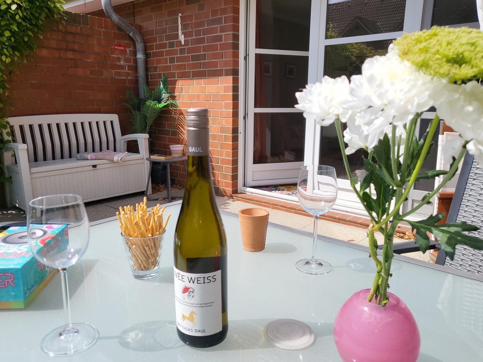 Außenbereich mit Tisch, Wein, Blumen und Gartenmöbeln. Hintergrund: rote Ziegelwand und Fenster.
