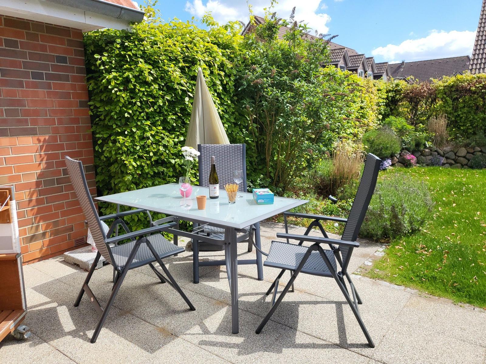 Terrasse mit Tisch und Stühlen, umgeben von grünen Hecken und Rasen.