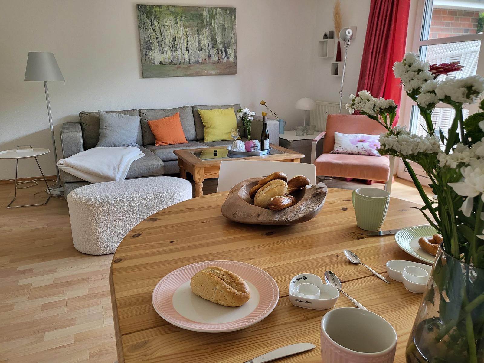 Ein Essbereich mit Holztisch, Brot und Blumen. Im Hintergrund ein Wohnzimmer mit Sofa und Kissen.