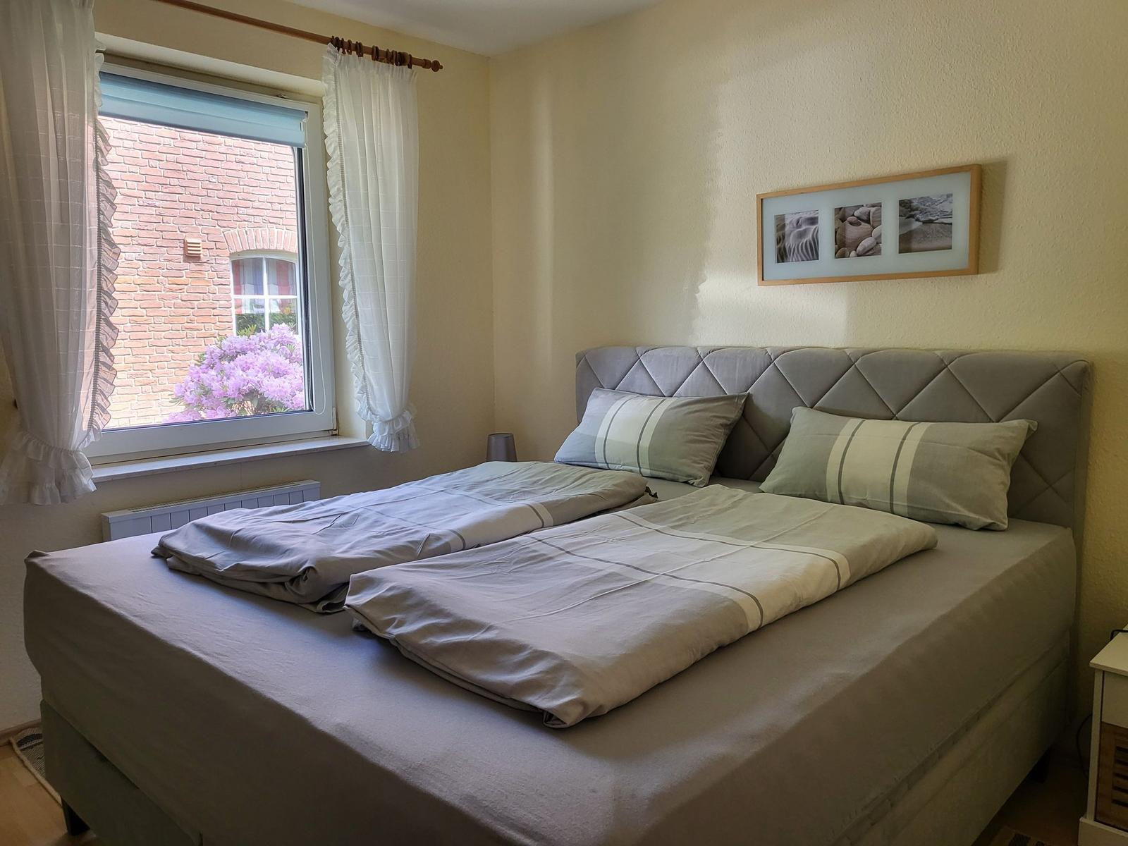 Schlafzimmer mit Doppelbett, Fenster mit Blick auf Haus und Blumen, Wandbild über dem Bett.