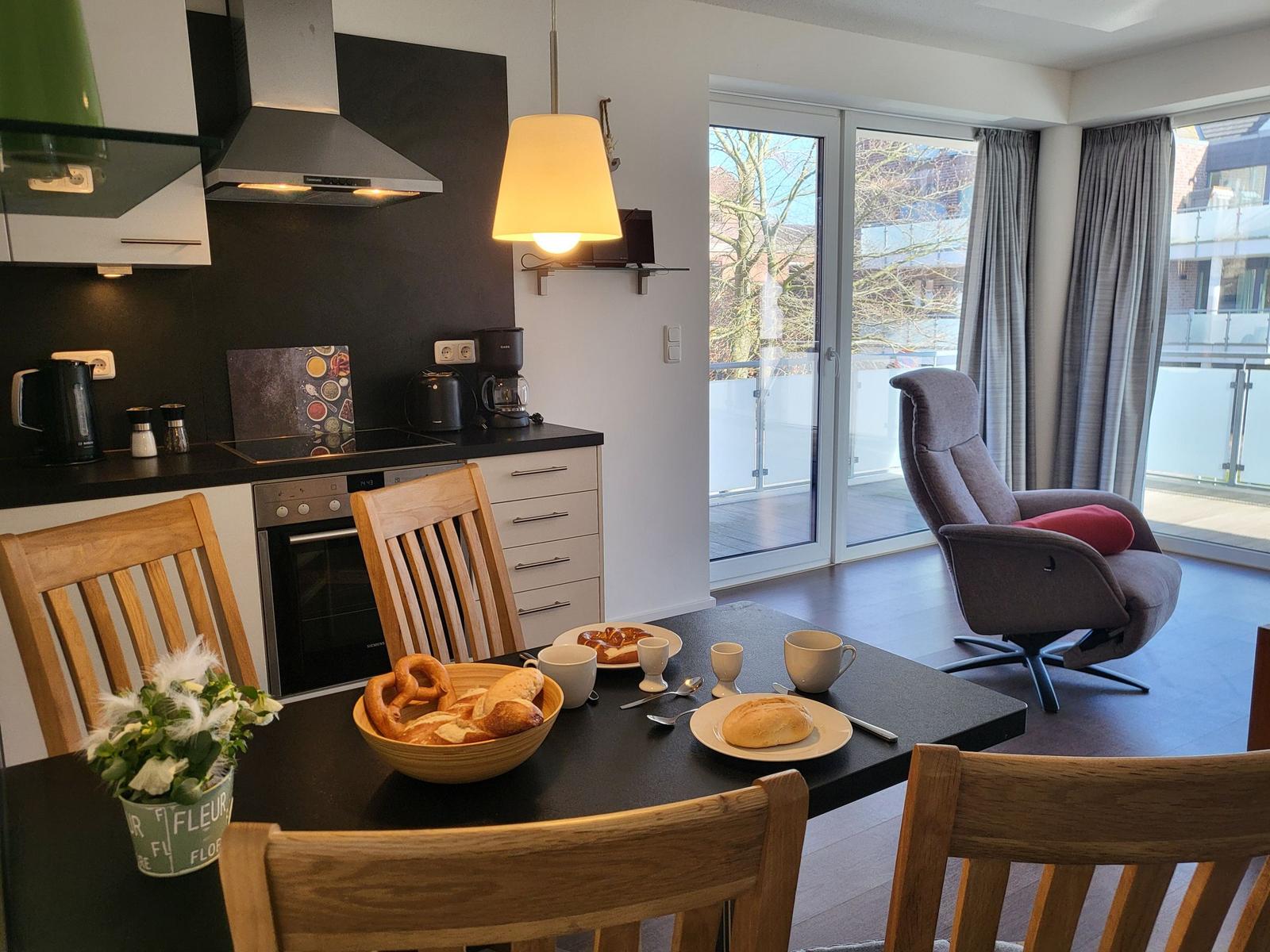 Essbereich mit Tisch, Stühlen und Blick auf Balkon. Küche mit Herd und Kaffeemaschine.