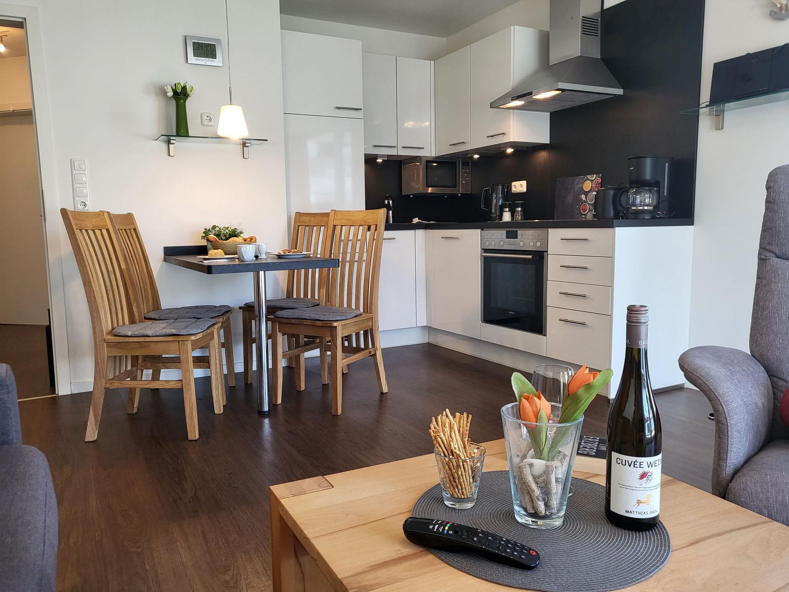 Küche mit Essbereich und Wohnzimmer. Holztisch, Stühle, Kühlschrank, Herd und Fernseher.