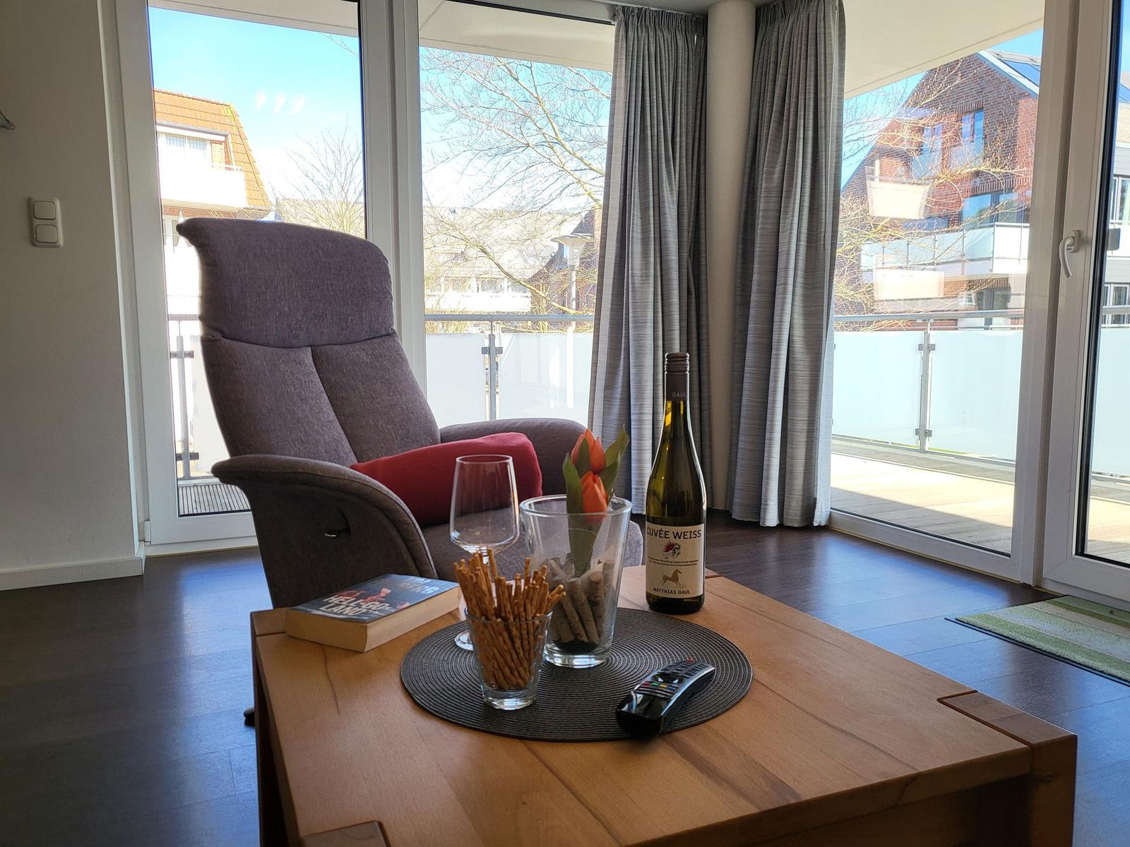 Ein Sessel mit rotem Kissen steht vor großen Fenstern. Auf dem Tisch stehen Wein, Tulpen und Snacks.