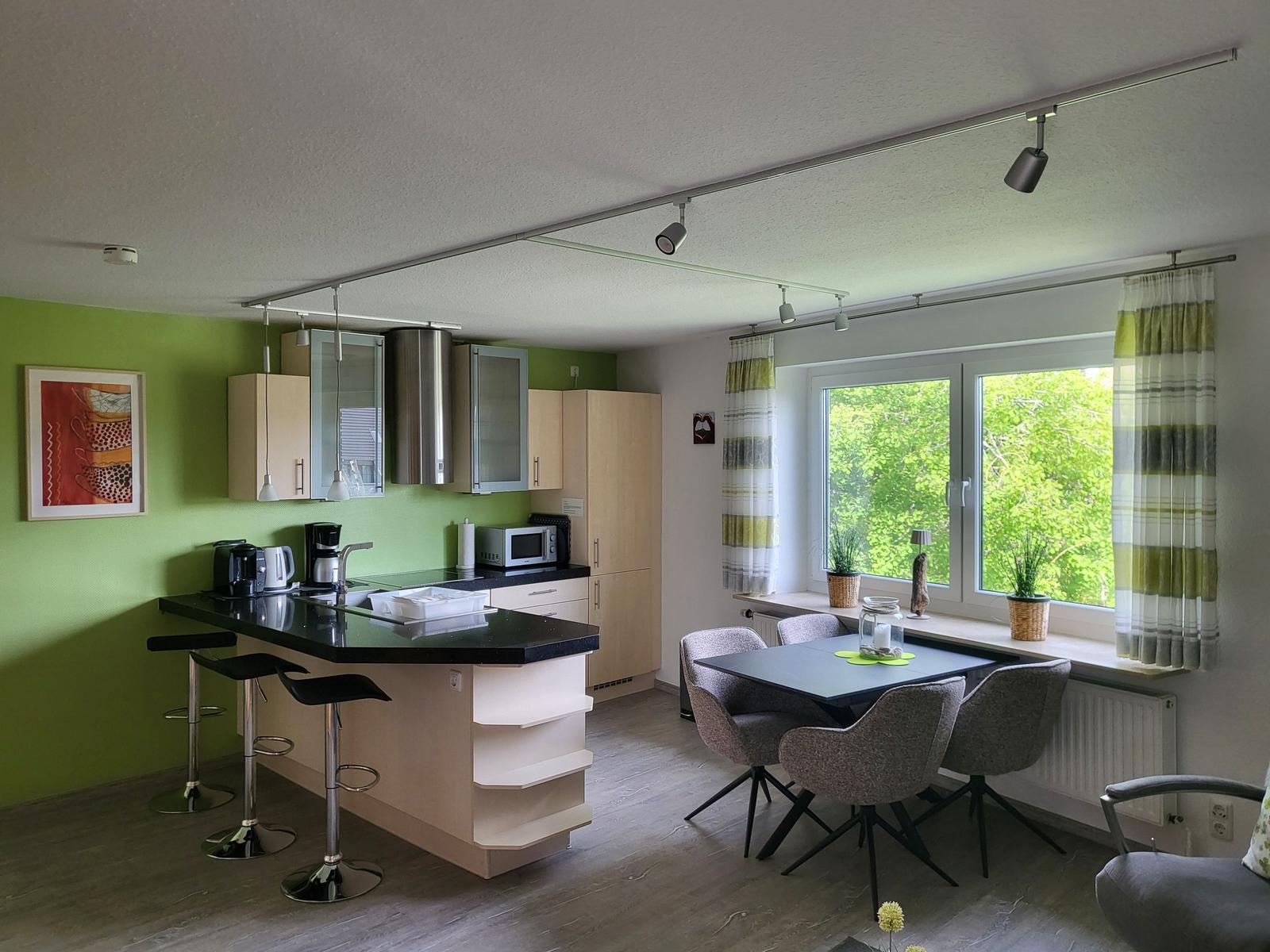Küche mit Essbereich und großem Fenster. Grüne Wand, schwarzer Arbeitsplatz.