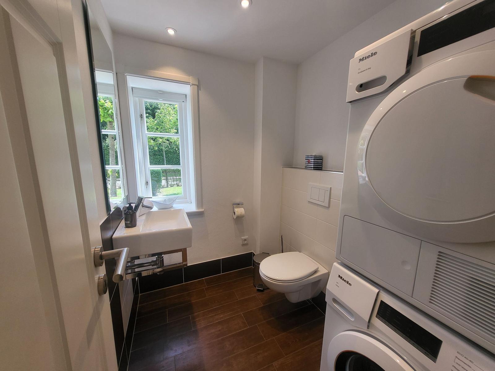 Badezimmer mit Waschmaschine, Trockner und Toilette. Fenster mit Blick auf Grün.